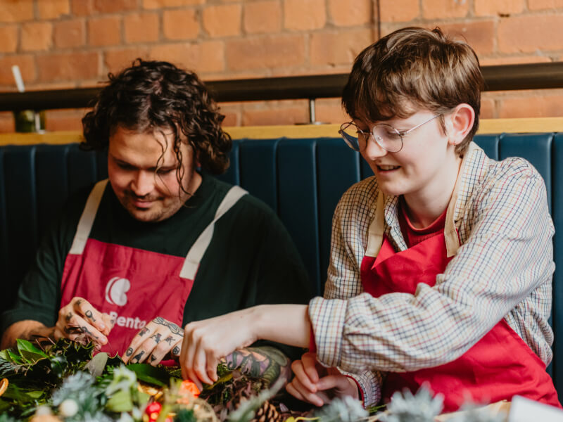Two people sitting at a table, making a festive wreath from a wreath making kit